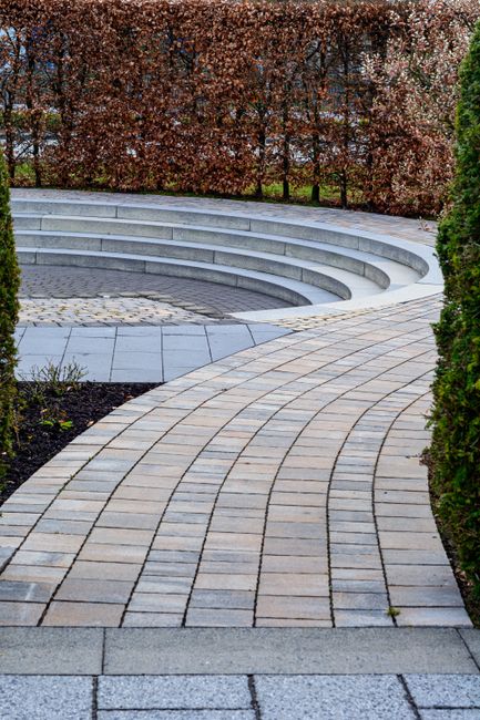 A paved garden pathway featuring curved, light-colored stone bricks that leads towards a low circular seating area with tiered steps made of similar grey paving stones. The path is bordered on one sid